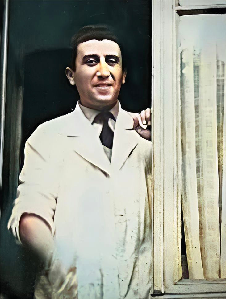 A man in a white coat stands by a window with sheer curtains. He has short dark hair and wears a dark tie. The setting appears to be indoors, and he is looking outward while resting one hand on the window frame.