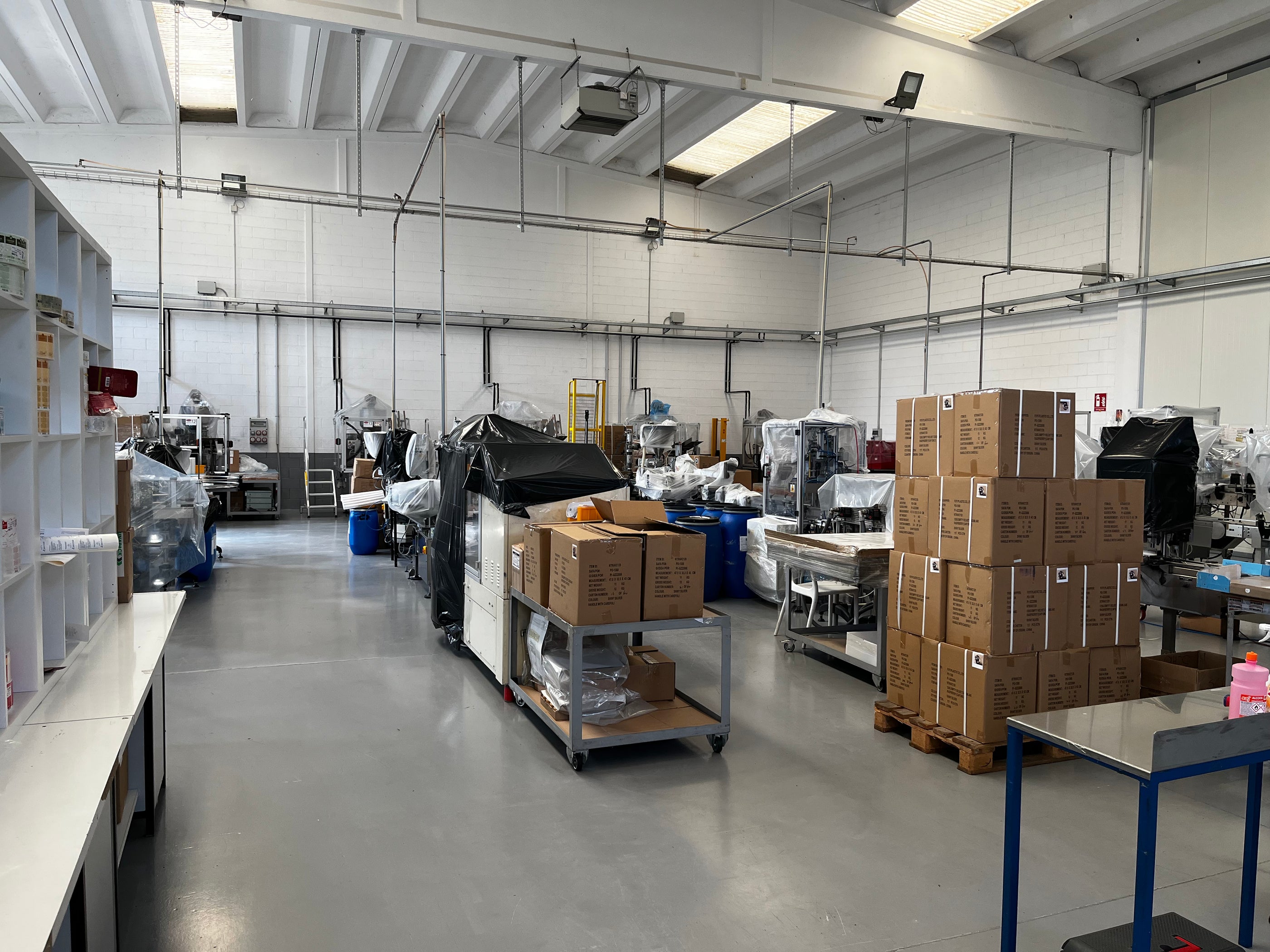 A spacious industrial warehouse with high ceilings, featuring stacks of cardboard boxes on the right and various packaging machines covered with plastic sheets throughout the area. The floor is concrete and shelving is visible on the left.